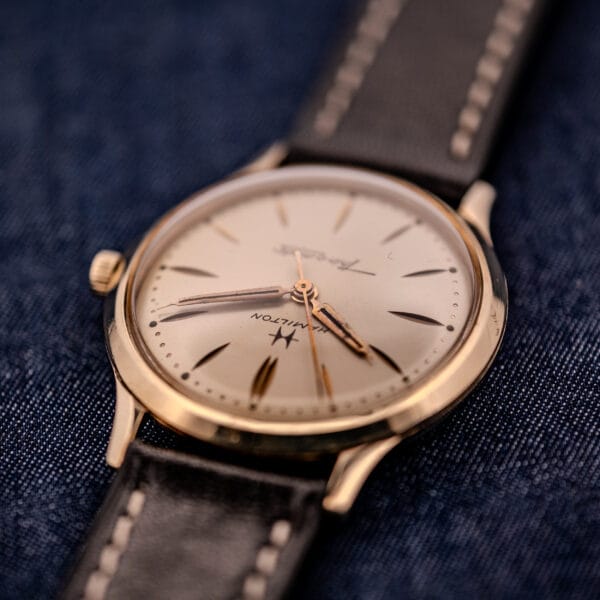 A 1962 Hamilton Thin-o-Matic Cal. 663 wristwatch with a 10K gold filled case and black leather strap rests on dark blue fabric.