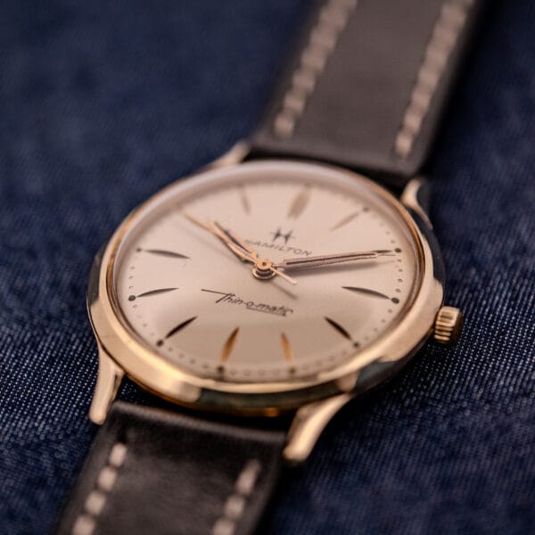 A 1962 Hamilton Thin-o-Matic Cal. 663 10K Gold Filled wristwatch with a black leather strap, displayed on dark blue fabric.