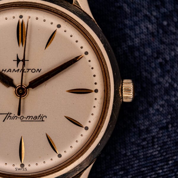 Close-up of a 1962 Hamilton Thin-o-Matic wristwatch with Cal. 663 movement, gold markers, and a 10K Gold Filled case, set against a textured dark blue background.