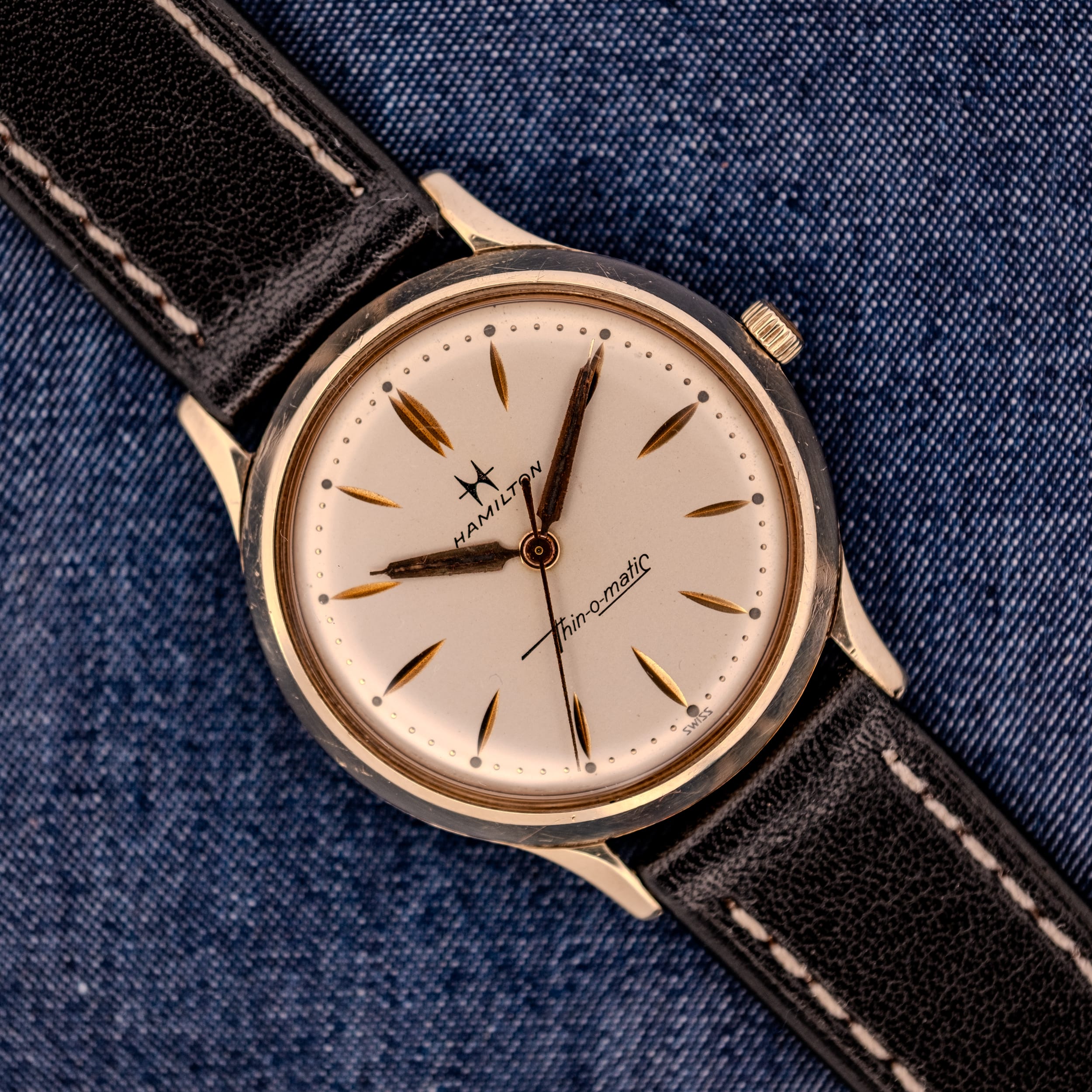 A 1962 Hamilton Thin-o-Matic Cal. 663 10K Gold Filled wristwatch with a black leather strap, displayed on denim fabric. A 1962 Hamilton Thin-o-Matic Cal. 663 10K Gold Filled wristwatch with a black leather strap, displayed on denim fabric.