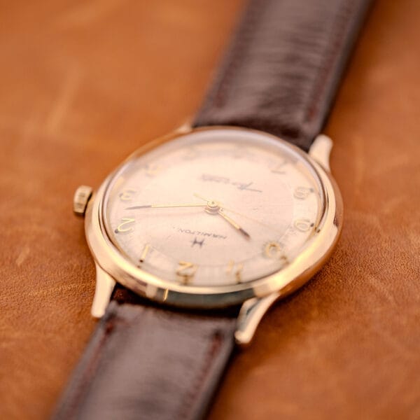 A 1966 Hamilton Thin-o-matic Cal. 668 10K Gold Filled wristwatch with a brown leather strap rests on a tan leather surface.