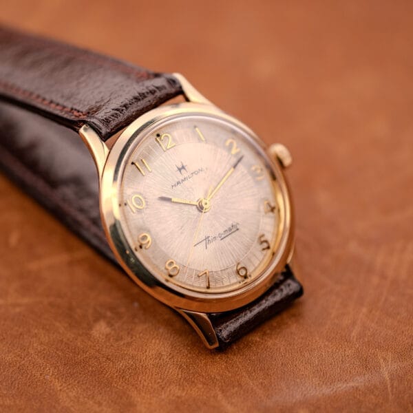 A 1966 Hamilton Thin-o-matic Cal. 668 10K Gold Filled wristwatch with a brown leather strap rests on a matching brown leather surface.