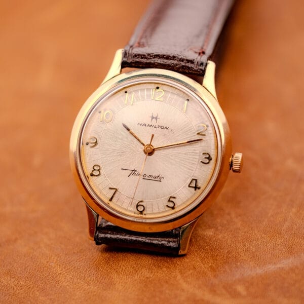 A 1966 Hamilton Thin-o-matic Cal. 668 10K Gold Filled wristwatch with leather strap is displayed on a brown surface.