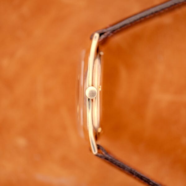 A close-up side view of a 1966 Hamilton Thin-o-matic Cal. 668 10K Gold Filled wristwatch with a brown leather strap on a brown background.