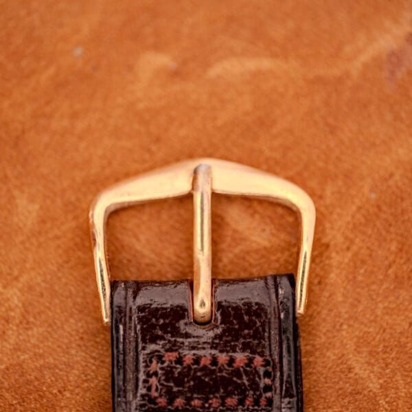 A close-up of the 1966 Hamilton Thin-o-matic Cal. 668 10K Gold Filled belt buckle on a dark brown leather strap, set against a tan background, highlighting its refined vintage style.