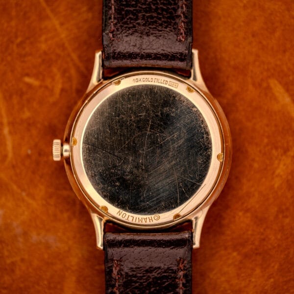A close-up of a 1966 Hamilton Thin-o-matic Cal. 668 10K Gold Filled watch back with a brown leather strap, displayed on a brown surface.