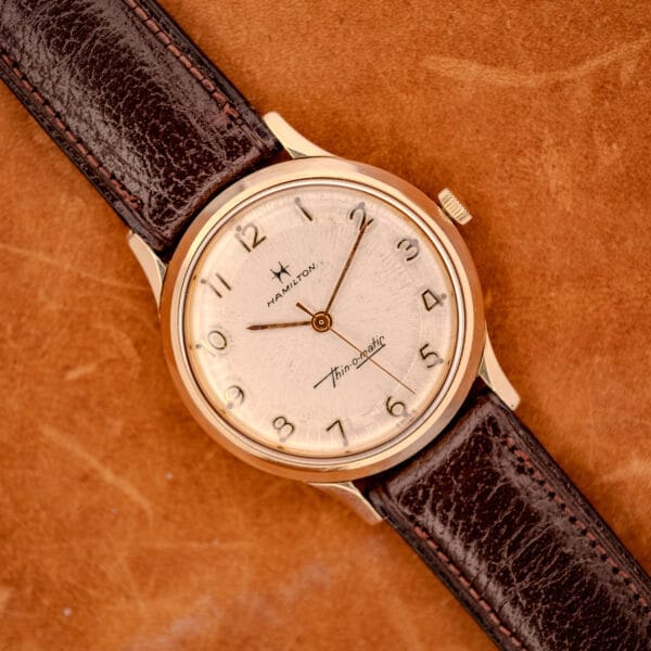 The 1966 Hamilton Thin-o-matic Cal. 668 10K Gold Filled is shown with a brown leather strap on a tan suede background.