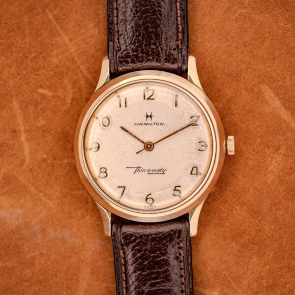 1966 Hamilton Thin-o-matic Cal. 668 wristwatch in 10K gold filled, paired with a brown leather strap and displayed on a tan leather background.