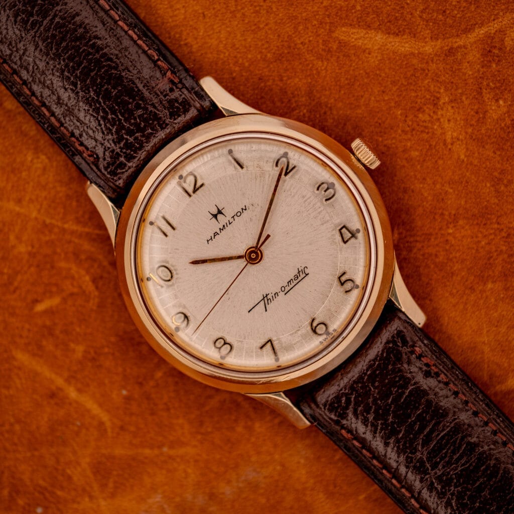A 1966 Hamilton Thin-o-matic Cal. 668 10K Gold Filled wristwatch with a brown leather strap rests elegantly on a brown surface.