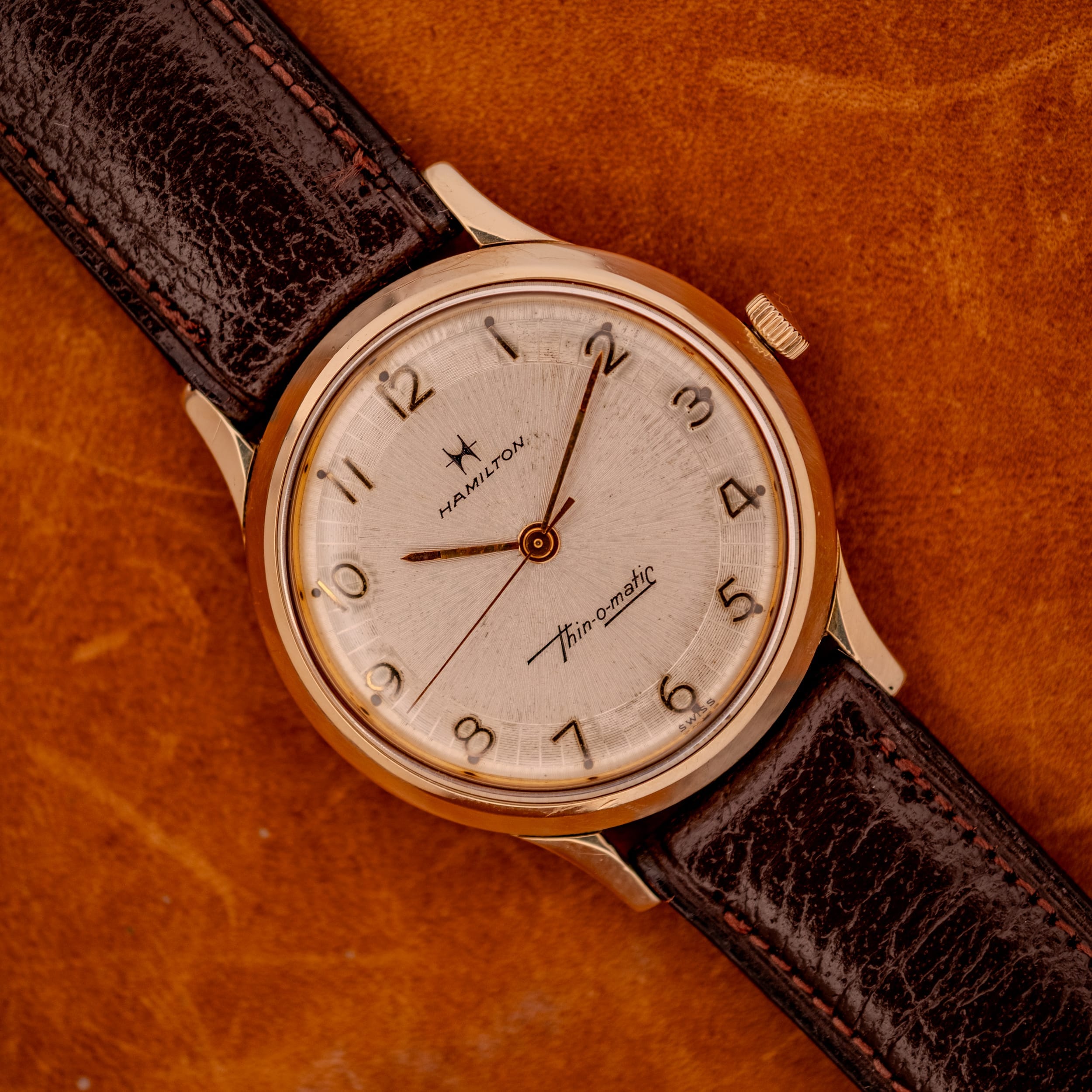 A 1966 Hamilton Thin-o-matic Cal. 668 10K Gold Filled wristwatch with a brown leather strap rests elegantly on a brown surface. A 1966 Hamilton Thin-o-matic Cal. 668 10K Gold Filled wristwatch with a brown leather strap rests elegantly on a brown surface.