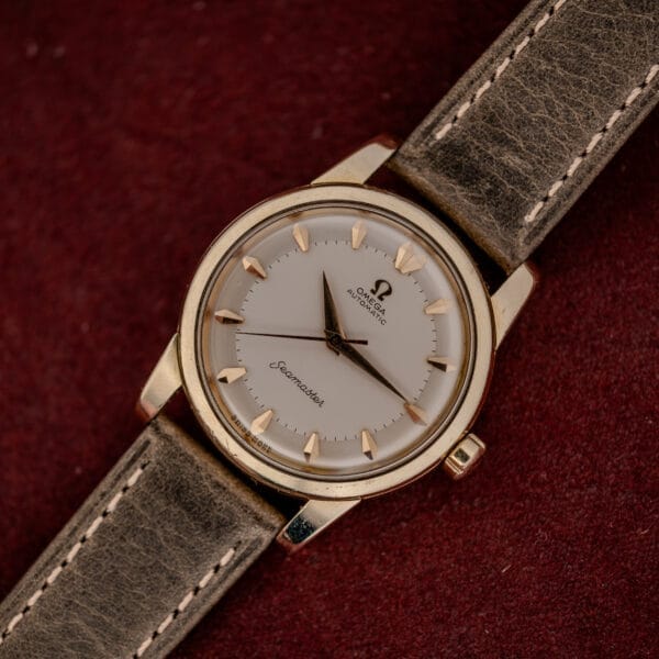 The Omega Automatic Seamaster Ref. 2846-2848 "Deluxe" features a gold capped case and brown leather strap, showcased against a red textured background.