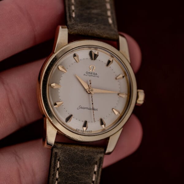 A hand holding an Omega Automatic Seamaster Ref. 2846-2848 "Deluxe" with a gold capped case and a brown leather strap.