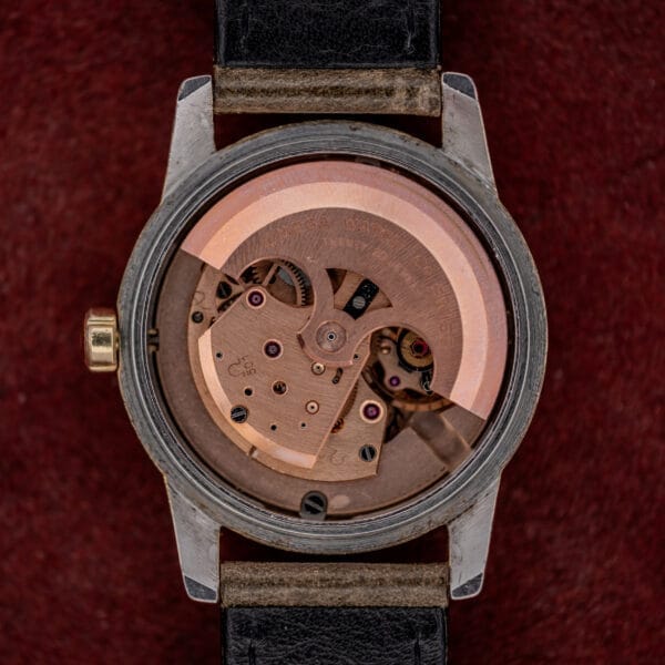 Close-up of the Omega Automatic Seamaster Ref. 2846-2848 "Deluxe" with exposed automatic movement, visible gears, black leather strap, and a luxurious gold capped case, set on a red surface.