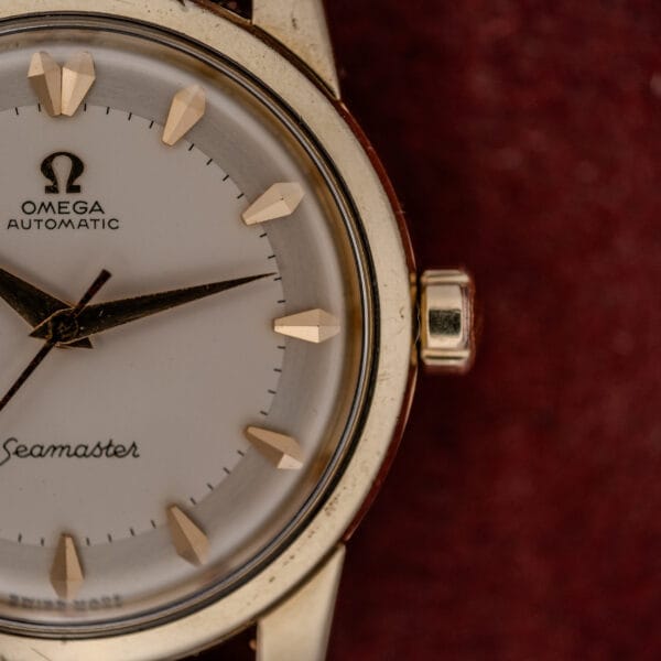 Close-up of an Omega Automatic Seamaster Ref. 2846-2848 "Deluxe" with a gold capped case and bezel, set against a maroon background.