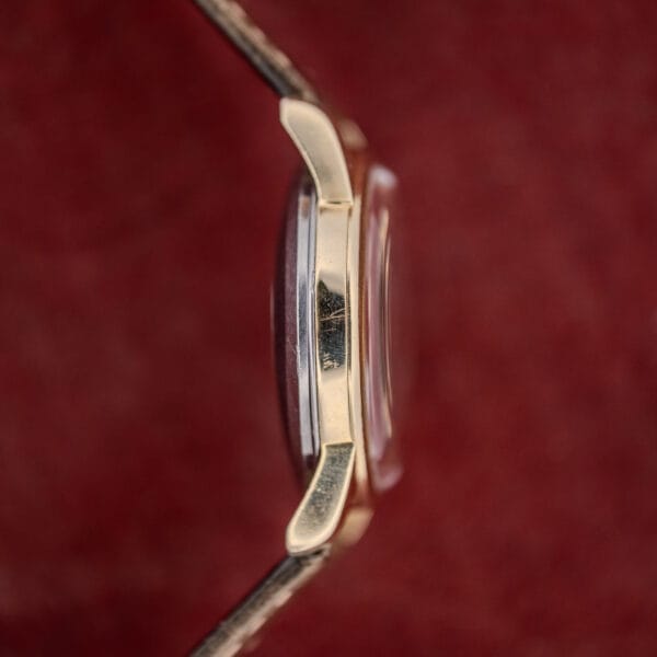 Side view of an Omega Automatic Seamaster Ref. 2846-2848 "Deluxe" with a gold capped case and clear crystal, set against a red textured background.