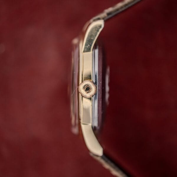 Close-up side view of the Omega Automatic Seamaster Ref. 2846-2848 "Deluxe" gold capped case crown against a blurred dark red background, highlighting its automatic movement.