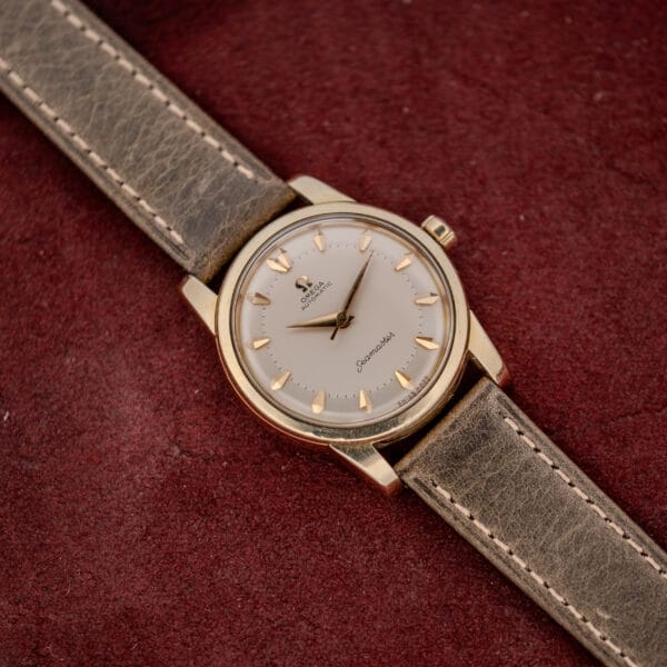 An Omega Automatic Seamaster Ref. 2846-2848 "Deluxe" with a gold capped case and beige leather strap is displayed on a red textured background.
