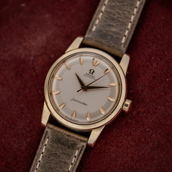 An Omega Automatic Seamaster Ref. 2846-2848 "Deluxe" with a gold capped case and tan leather strap sits on a burgundy textured surface.