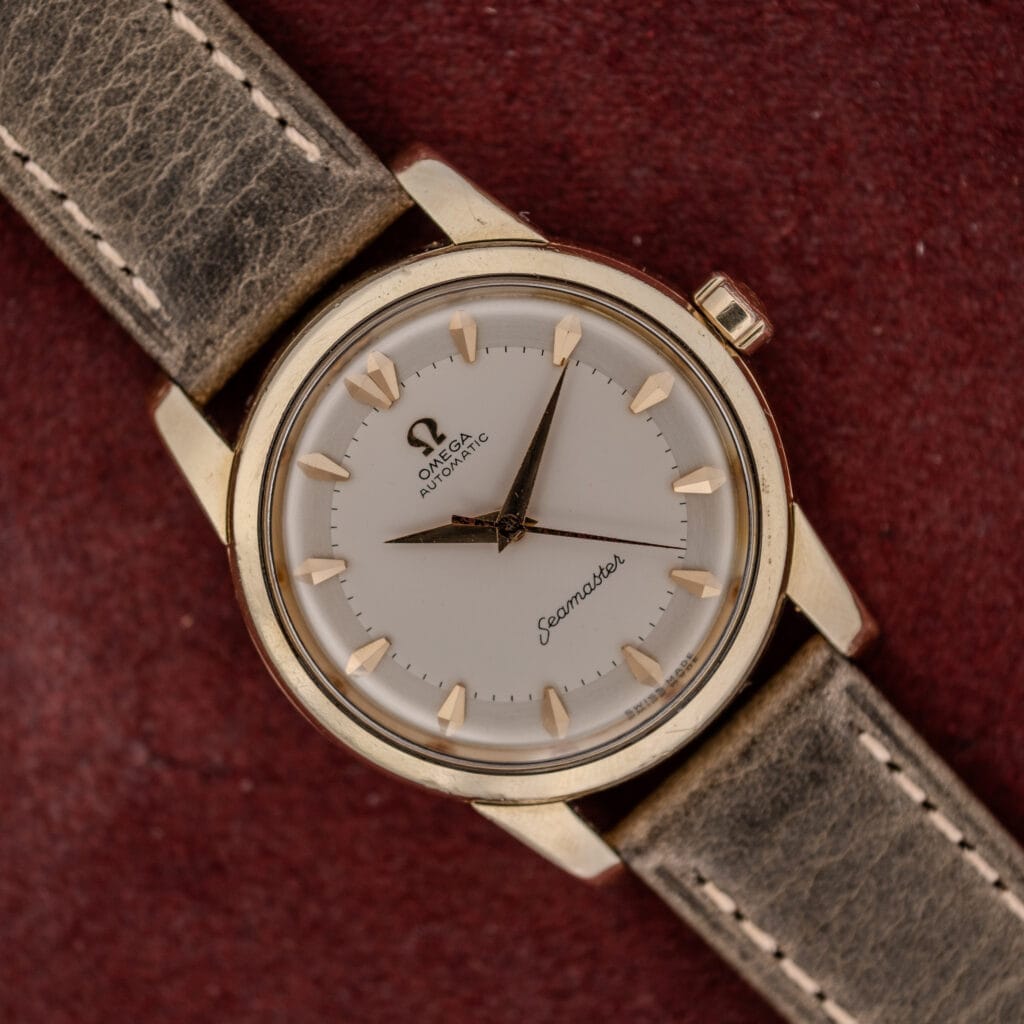An Omega Automatic Seamaster Ref. 2846-2848 "Deluxe" with a gold capped case and worn leather strap rests elegantly on a maroon background.