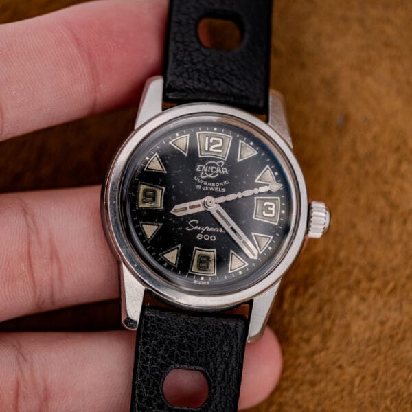 A hand holding a 1967 Enicar Seapearl 600 Sherpa Ref. 100/120 Cal. 1010 stainless steel wristwatch with a black dial and leather strap.