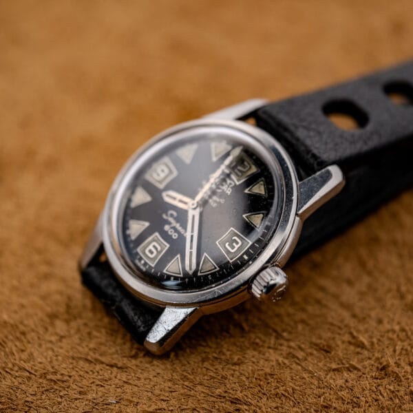 A 1967 Enicar Seapearl 600 Sherpa Ref. 100/120 Cal. 1010 stainless steel wristwatch with a black strap is displayed on a brown textured surface.