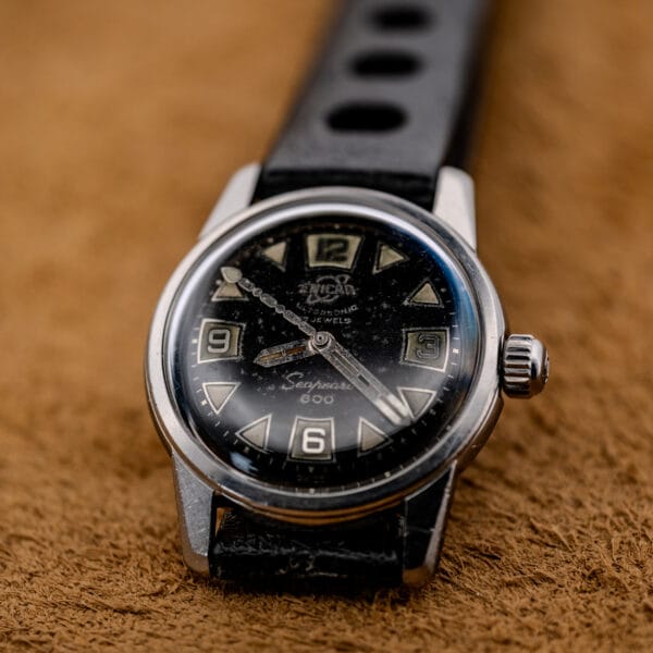 Close-up of a 1967 Enicar Seapearl 600 Sherpa Ref. 100/120 Cal. 1010 stainless steel wristwatch with black dial and leather strap on a brown textured surface.