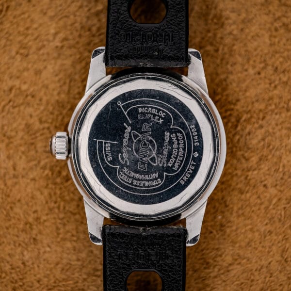 Close-up of the back of a 1967 Enicar Seapearl 600 Sherpa Ref. 100/120 Cal. 1010 Stainless Steel watch with a black strap, resting on a brown textured surface.