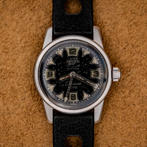A 1967 Enicar Seapearl 600 Sherpa Ref. 100/120 Cal. 1010 stainless steel wristwatch with a black dial and black leather strap is displayed on a brown textured background.