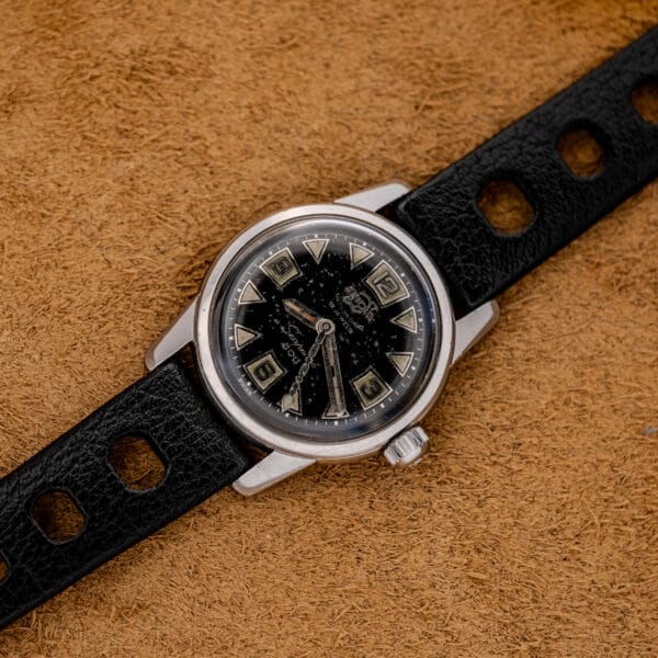 A 1967 Enicar Seapearl 600 Sherpa Ref. 100/120 Cal. 1010 stainless steel wristwatch with a perforated black leather strap rests on a textured brown surface.