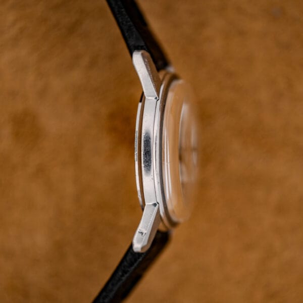 Side view of a 1967 Enicar Seapearl 600 Sherpa Ref. 100/120 Cal. 1010 Stainless Steel wristwatch with a black strap and silver case, set against a brown textured background.