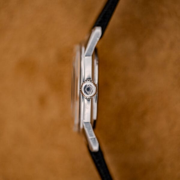Side view of a 1967 Enicar Seapearl 600 Sherpa Ref. 100/120 Cal. 1010 Stainless Steel wristwatch with a black strap, set against a blurred brown background.