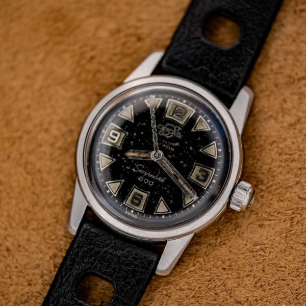 A vintage wristwatch with a black dial and bold numerals on a black leather strap, set on brown fabric—this timepiece mirrors the classic style of the 1967 Enicar Seapearl 600 Sherpa Ref. 100/120 Cal. 1010 Stainless Steel.