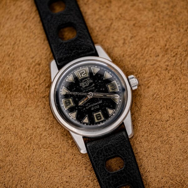 A 1967 Enicar Seapearl 600 Sherpa Ref. 100/120 Cal. 1010 stainless steel wristwatch with a black leather strap and black dial, displayed on a brown textured surface.