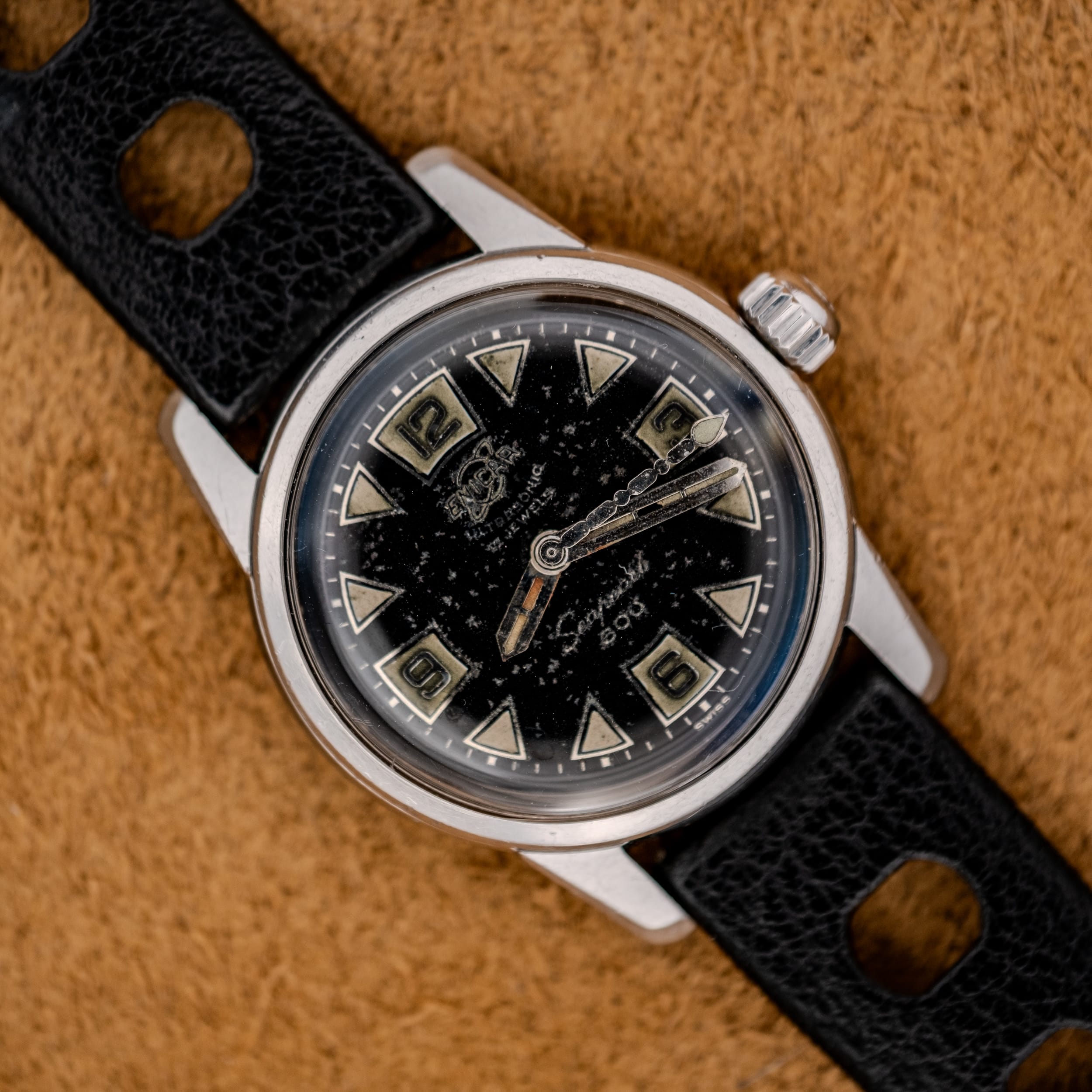 A 1967 Enicar Seapearl 600 Sherpa Ref. 100/120 Cal. 1010 stainless steel watch with a black leather strap rests elegantly on a textured brown surface. A 1967 Enicar Seapearl 600 Sherpa Ref. 100/120 Cal. 1010 stainless steel watch with a black leather strap rests elegantly on a textured brown surface.