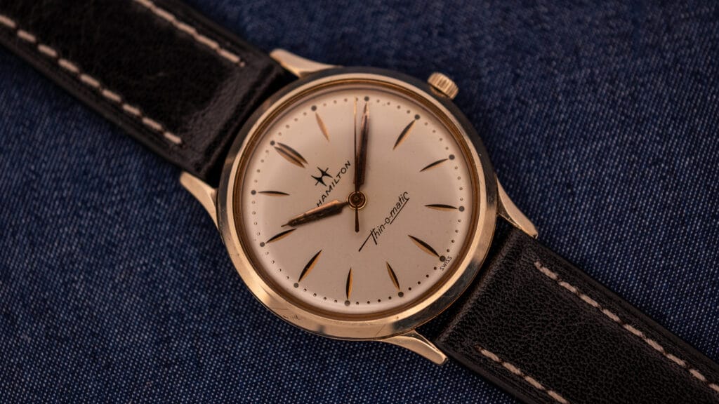 A vintage Hamilton Thin-O-Matic wristwatch with a black leather strap rests elegantly on blue fabric, showcasing the classic style and precision of Hamilton craftsmanship.