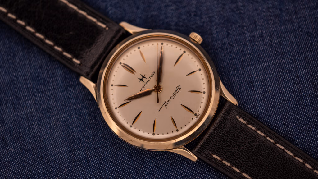A vintage Hamilton Thin-O-Matic wristwatch with a black leather strap rests elegantly on blue fabric, showcasing the classic style and precision of Hamilton craftsmanship.