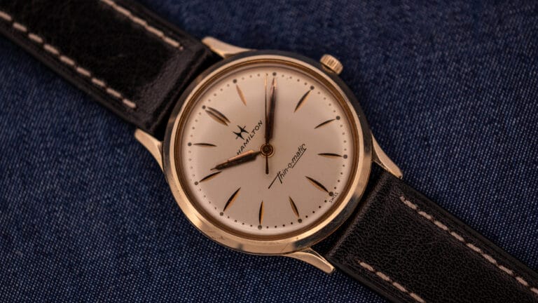A vintage Hamilton Thin-O-Matic wristwatch with a black leather strap rests elegantly on blue fabric, showcasing the classic style and precision of Hamilton craftsmanship.