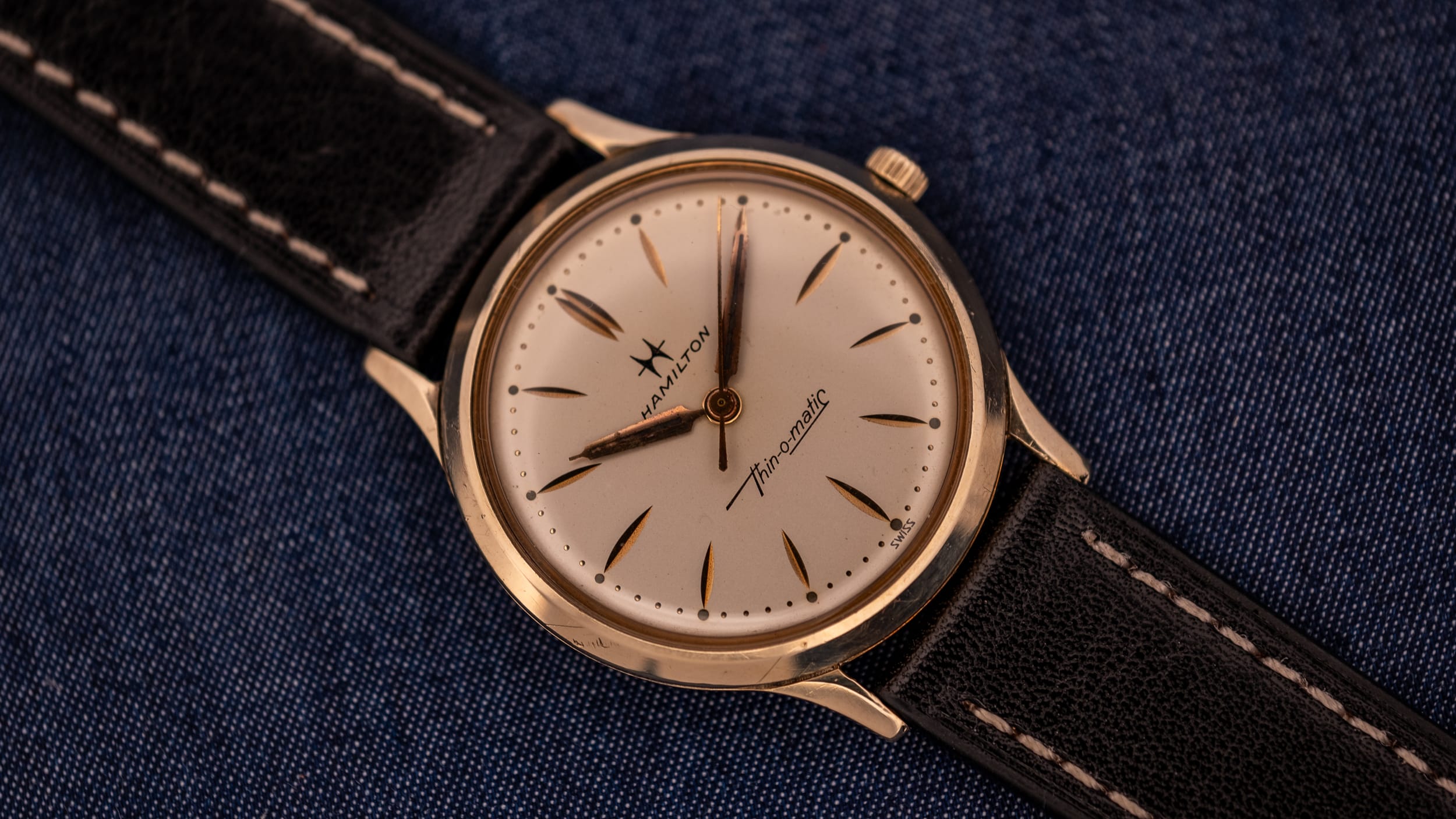 A vintage Hamilton Thin-O-Matic wristwatch with a black leather strap rests elegantly on blue fabric, showcasing the classic style and precision of Hamilton craftsmanship.