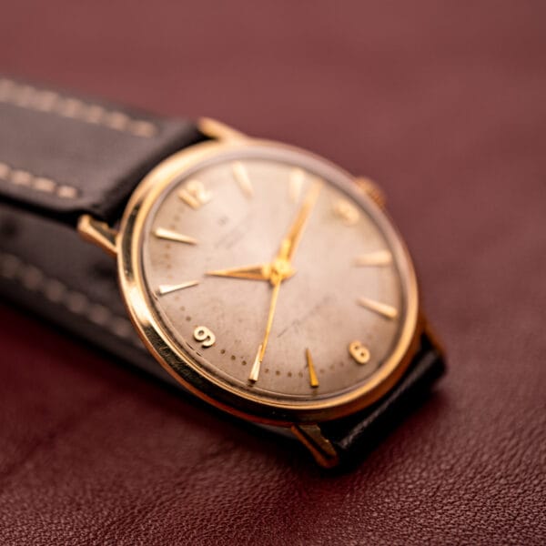 A Hamilton Thin-O-Matic 10k Gold Filled Case Automatic wristwatch with a leather strap is elegantly displayed on a brown textured surface.