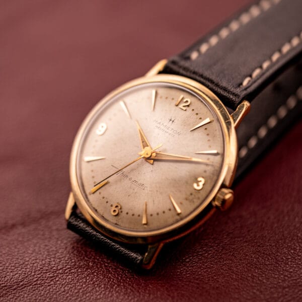 A Hamilton Thin-O-Matic 10k Gold Filled Case Automatic wristwatch with a black leather strap is displayed elegantly on a maroon surface.