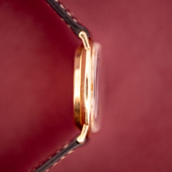 Side view of a Hamilton Thin-O-Matic 10k Gold Filled Case Automatic wristwatch with black leather strap, shown against a blurred red backdrop.