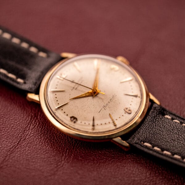 Close-up of a Hamilton Thin-O-Matic 10k Gold Filled Case Automatic wristwatch, featuring a gold filled case and automatic movement with a black leather strap, displayed on a maroon surface.