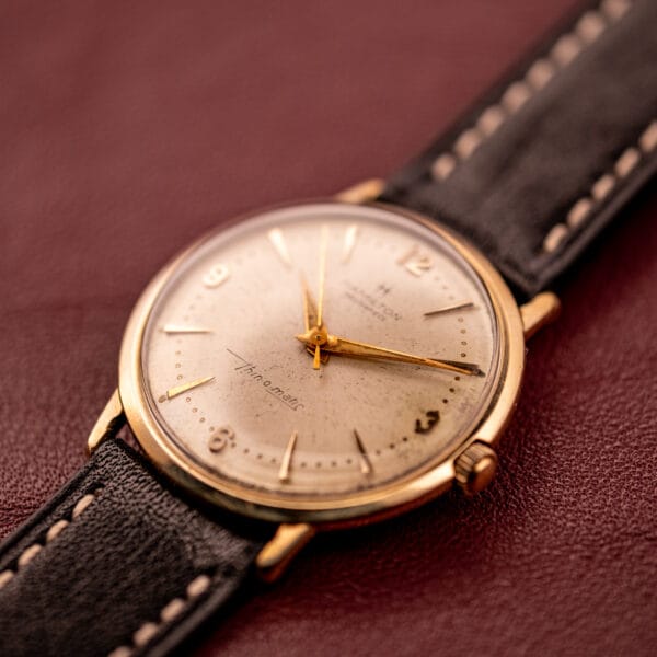 The Hamilton Thin-O-Matic 10k Gold Filled Case Automatic wristwatch with a black leather strap rests on a brown surface, displaying the time 10:08.