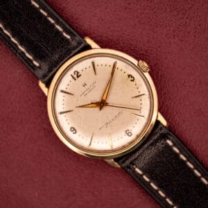 The Hamilton Thin-O-Matic 10k Gold Filled Case Automatic watch with a black leather strap is displayed on a maroon background.