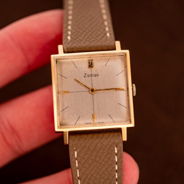A hand holds a Zodiac Ref. 523-007 Square Case Manual Wind Cal. 52 wristwatch featuring gold accents and a brown leather strap.