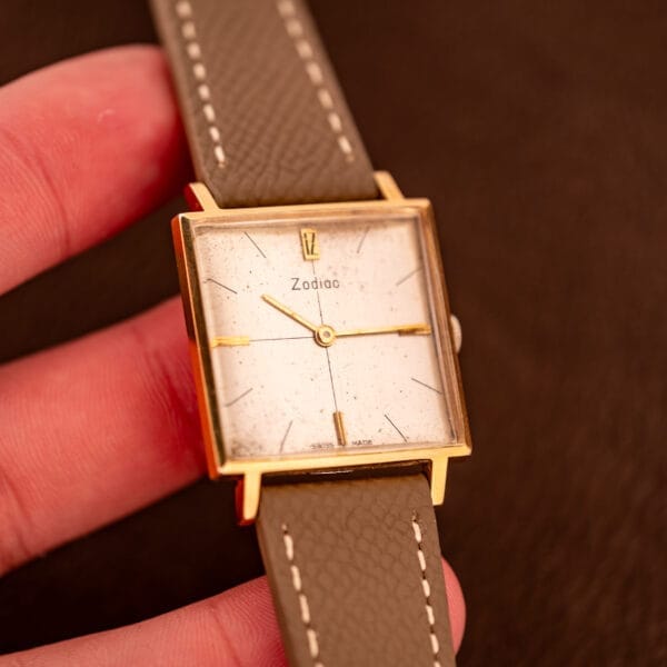A hand holding a Zodiac Ref. 523-007 wristwatch featuring a square gold case, white dial, manual wind Cal. 52 movement, and a light brown leather strap.