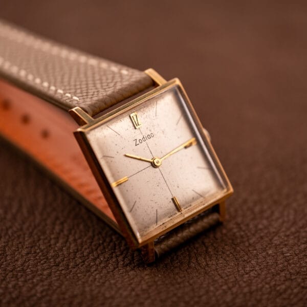The Zodiac Ref. 523-007, featuring a square case and brown leather strap, rests on a textured brown surface. This vintage timepiece is powered by manual wind Cal. 52 movement.