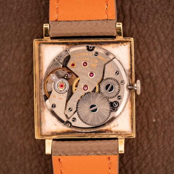 The Zodiac Ref. 523-007 features a square gold case with visible manual wind Cal. 52 movement and is paired with a brown leather strap, shown on a textured surface.
