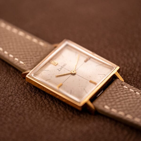A Zodiac Ref. 523-007 Square Case Manual Wind Cal. 52 wristwatch with a brown leather strap sits on a textured brown surface.