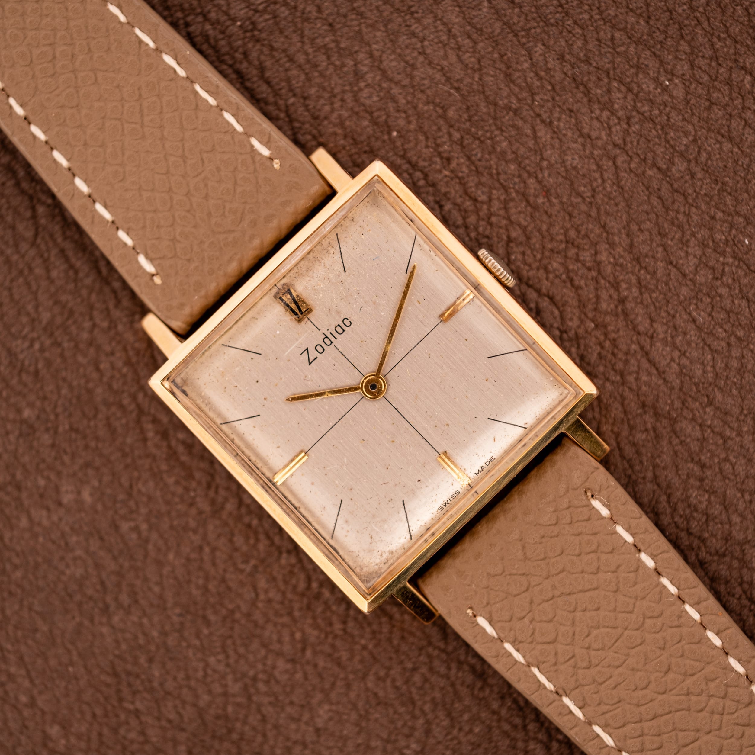 A Zodiac Ref. 523-007 Square Case Manual Wind Cal. 52 wristwatch with a tan leather strap rests on a brown textured surface. A Zodiac Ref. 523-007 Square Case Manual Wind Cal. 52 wristwatch with a tan leather strap rests on a brown textured surface.
